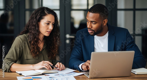 Two diverse colleagues collaborating on a project, reviewing documents and using a laptop in an office setting