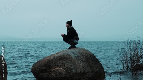 A lonely girl is sitting on a big stone on the shore of a lake on a cloudy rainy day and enjoying nature. Cool weather. The harmony of man and nature.