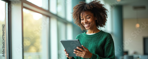 Happy black woman uses tablet. Smiling african woman looks at tablet screen in office or home. Lady wearing green sweater uses portable device. She enjoys tech. Window behind her. © Vadym
