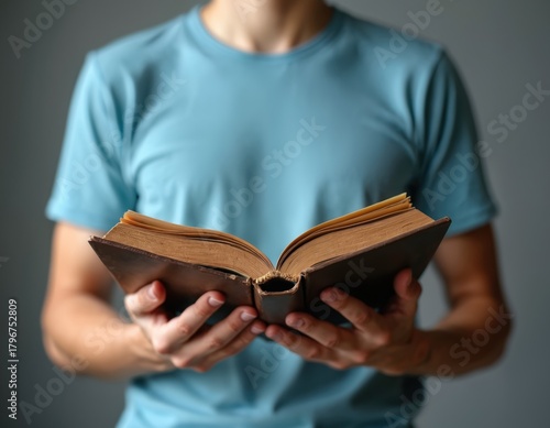 Young man in blue shirt holds open old book with both hands. He reads a vintage book, searching for knowledge and wisdom. He learns, studies, gains info from classic literature and education.