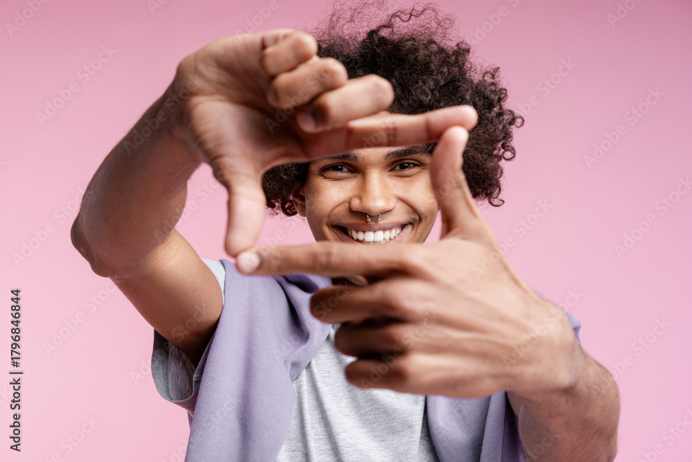 Fototapeta premium Smiling happy Latin man with piercing in nose showing frame with fingers isolated on pink background