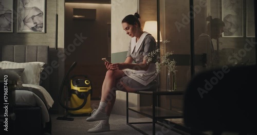 Wallpaper Mural Tired Housekeeping Worker Pauses to Breathe After Cleaning. Maid in Professional Uniform Sits Quietly in Hotel Room Armchair, Scrolling Phone With Calm Expression. Concept Self Care at Work. Handheld. Torontodigital.ca