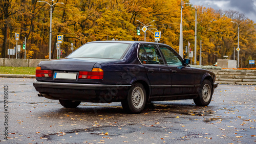 Принти на полотні Blue classic sedan parked on a rainy autumn lot, set against wet trees with yell