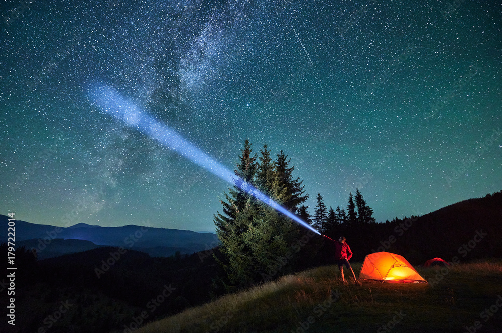 Obraz premium Man hiker stands under dazzling starry sky, shining flashlight into heavens. Milky Way stretches above, glowing tent nearby, creating magical and serene scene in peaceful mountain wilderness.