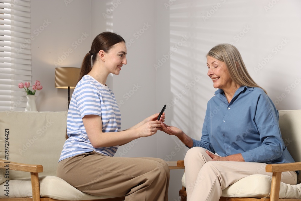 Obraz premium Diabetes. Smiling daughter checking mother's blood sugar level with lancet pen on armchair at home