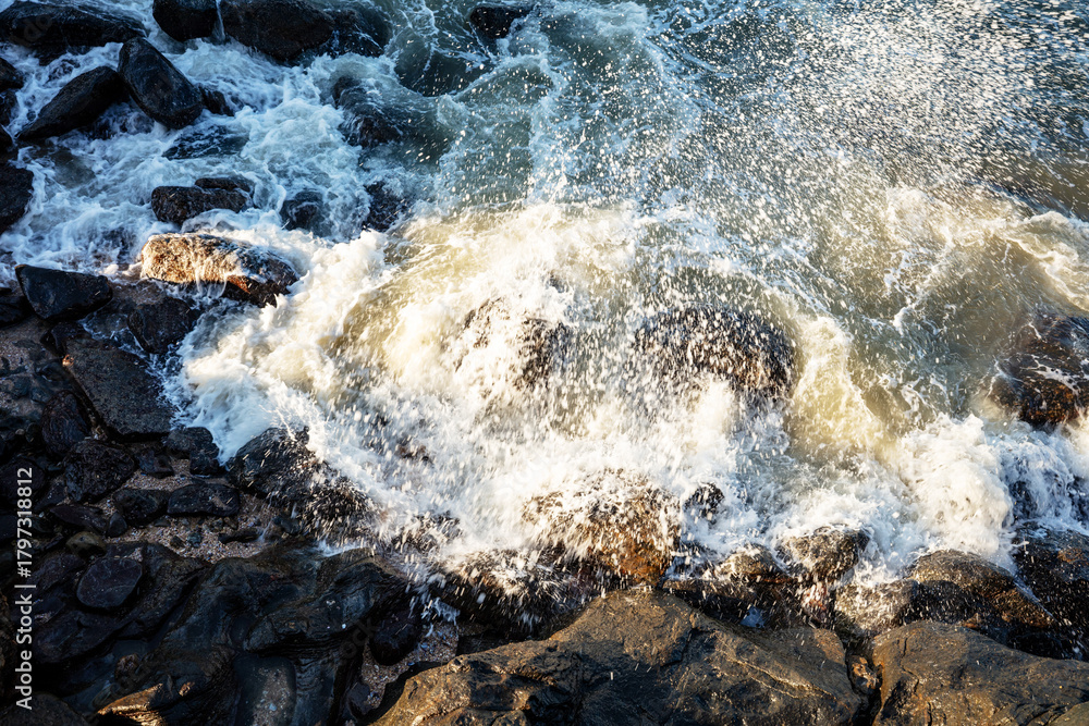 Obraz premium Ocean waves crashing against the beach rocks, light reflecting on the water.