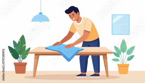 Cheerful young man in a yellow t-shirt and blue pants diligently wiping a wooden table with a blue cloth, performing household chores with a smile.
