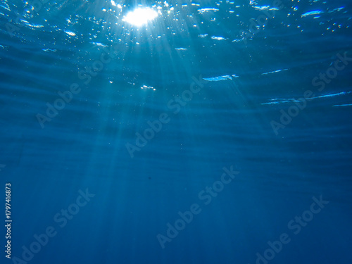 Dark blue ocean surface seen from underwater. Abstract waves underwater and rays of sunlight shining through, Sun light rays undersea deep, Underwater background with sea bottom, Mediterranean sea.