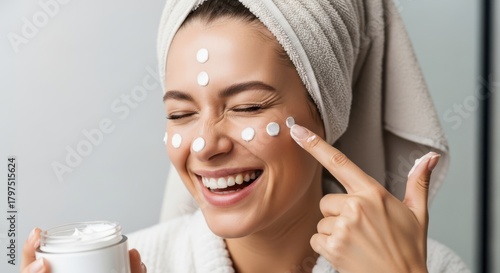 A happy woman with a towel on her head applies cream to her face, smiling with her eyes closed, holding a jar of moisturizer
