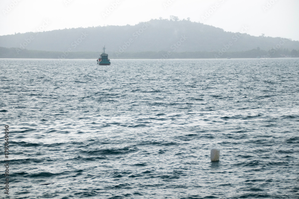 Obraz premium A solitary boat on a misty sea with the silhouette of a mountain in the background during a monsoon rain. Koh Sdach in Sihanoukville, Cambodia