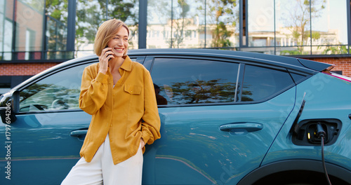 Beautiful woman in casual clothes talking on the phone with a friend while standing next to her electric car charging at a portable charging station on a city street.