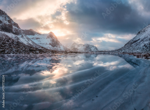 Aerial view of frozen lake in Lofoten, Norway, snow-covered mountains and low winter sun reflecting on the icy surface at sunset. Cold Arctic landscape, dramatic clouds. Winter scenery. Top drone view