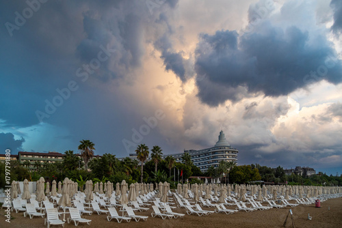 Fototapeta Naklejka Na Ścianę i Meble -  Empty white sunbeds and closed umbrellas arranged in neat rows on a gloomy beach day, with hotels and palm trees under low rain clouds with diffused sunlight. Side, Antalya, Turkey.

