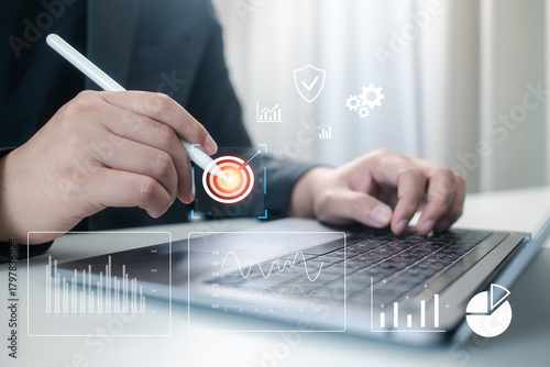 A professional uses a digital pen to target a goal on a holographic data interface above a laptop. This image symbolizes strategy, security, business intelligence, and performance measurement.