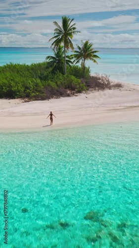 Aerial view of a young woman walking along a pristine white sand beach on a tropical island, surrounded by crystal-clear turquoise water and lush palm trees
