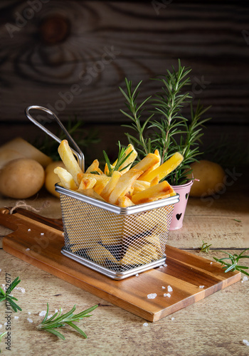 Golden french fries in metal basket with rosemary on wood
