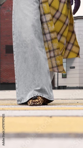 Close up of stylish woman crossing street wearing wide leg jeans, yellow plaid coat and leopard print shoes. Trendy street fashion detail in city outdoors