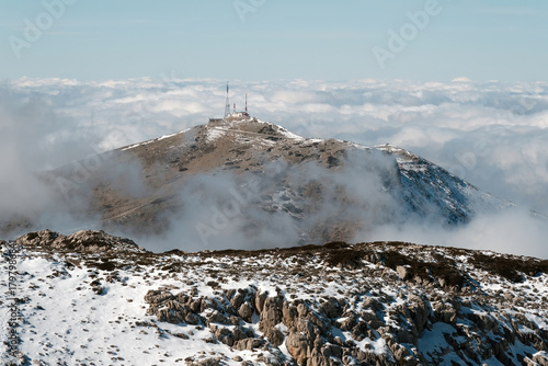 A snow-dusted peak in the Sierra de Segura rises through low clouds, with a communication station visible at the summit under a pale blue sky.