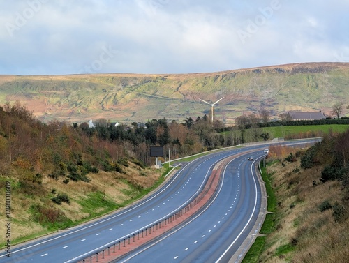 An elevated wide shot captures a modern, dual-carriageway highway curving through a rolling, semi-rural landscape under a pale, overcast sky. The road is flanked by grassy slopes and sparse trees