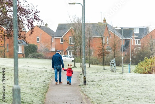 Father walking a toddler child hand in hand along a frosty paved path in a suburban area with houses and trees.