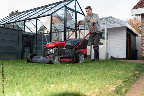 Man mowing narrow backyard lawn beside modern metal fence and garden plants.