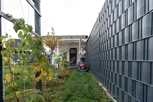 Man mowing narrow backyard lawn beside modern metal fence and garden plants.