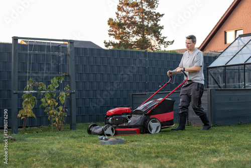 Man mowing lawn with red mower in backyard garden on sunny afternoon.