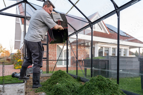 Man empties grass clippings into greenhouse for composting near modern brick house.