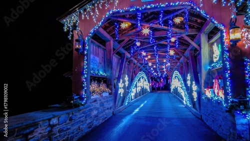 Colorful lights hang from the wooden beams of a covered bridge. The scene is filled with winter decorations, creating a joyful and magical atmosphere during the evening hours of a holiday festival.