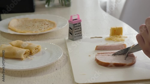 Hands slicing ham on cutting board with plate of rolled crepes and mini grater, soft morning light, home cook preparing simple breakfast, close-up of knife movement and textured food details