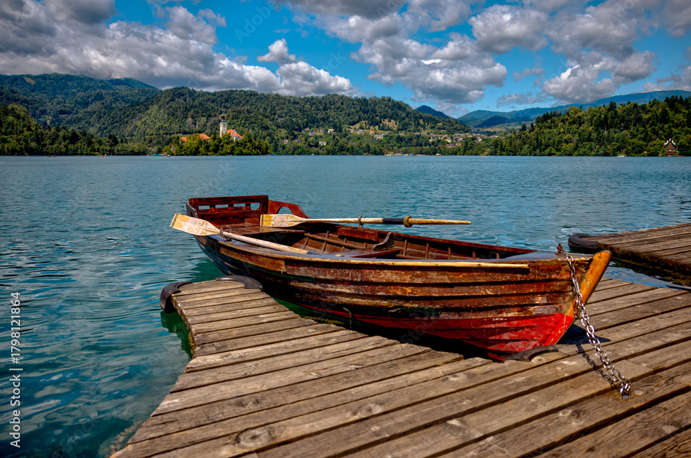 Obraz premium Traditional wooden pletna boats docked along the calm waters of Lake Bled, Slovenia, surrounded by lush greenery and the Julian Alps in the distance on a serene summer morning. 