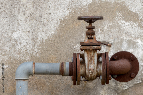 Old rusty industrial faucet, outdoor, with rusty pipes disappearing into the wall in the background.