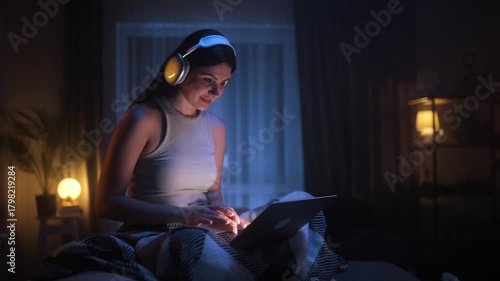 Young woman using laptop at night with headphones on, sitting under a blanket in cozy dim light. Relaxed evening mood, watching videos or chatting online