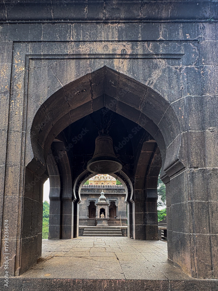 Naklejka premium A serene view of an ancient temple at Wai, Maharashtra, framed through stone arches with a large bell and intricate carvings, showcasing rich Maratha-era architecture.