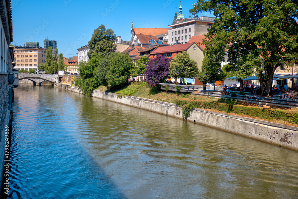 Obraz premium Ljubljana, Slovenia – July 14, 2025: A scenic view of the Ljubljanica River as it winds through the historic center of Ljubljana. Lined with trees, cafés, and charming riverside architecture. 