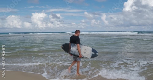 A man walks into the ocean with a black surfboard.