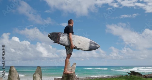 Surfer with black surfboard standing on a wooden stump looking at the waves in ocean. 