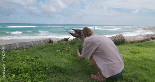 Woman takes a picture or video of flowers by the beach with ocean waves in the background for social media.