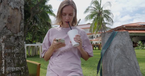 In a tropical hotel, a woman engaged with her smartphone. She drinks refreshing drink, enjoying the relaxed atmosphere and sunny weather.