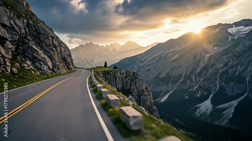A winding road through mountainous terrain, with sun setting over distant peaks. Clouds and golden light