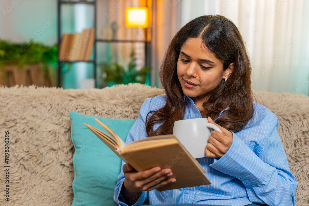 Fototapeta premium Indian young woman enjoys morning coffee while reading book on home sofa, feeling calm and thoughtful. Arabian girl embraces peaceful moment with warm drink and quiet literary focus in early hours.