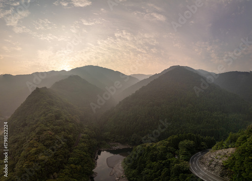 A stunning aerial view of a mountain range at sunrise, with a winding road cutting through a lush valley and a river flowing through it. The sky is filled with dramatic clouds.