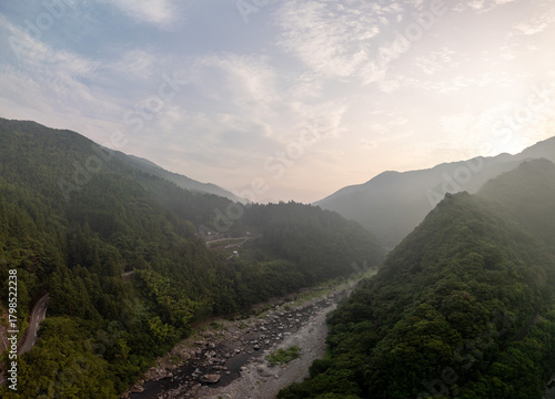 A stunning aerial view of a lush, green valley with a winding river and towering mountains under a cloudy sky at sunrise.