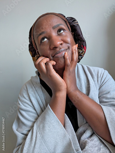 Foto A young African American woman in a robe and bonnet, gently holding her face while looking up with a soft, amused expression