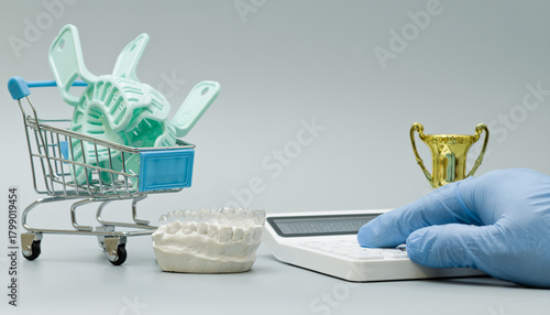 Mini shopping cart filled with dental impression trays beside a jaw model, calculator, and trophy, symbolizing cost, quality, and dental excellence.