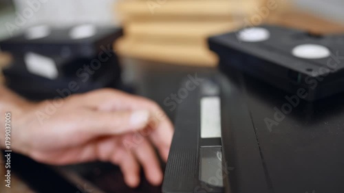 VHS cassette on VCR surface. A videotape rests partly inserted into a VCR with a blurred television behind it.