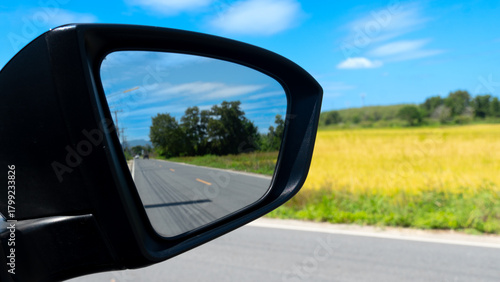 Car side mirror with shadow of asphalt road in the mirror. Travel of car on the aspahlt road with view of the golden rice fields. Rural agricultural areas of Thailand.