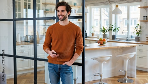 Cheerful man in sweater holding a phone standing in a modern apartment with kitchen view