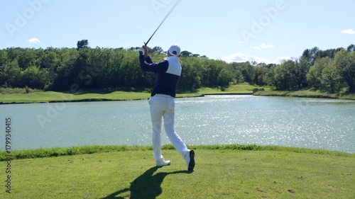 Golfer swings club by the water on a sunny day in the park
