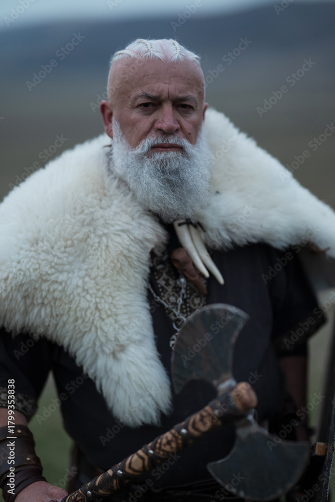Obraz premium Viking warrior with axe standing on mountain cliff, wearing fur cloak and leather armor, overlooking dramatic landscape. Historical Nordic fighter